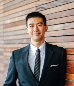 Man in suit smiling, leaning against wood panel. Volunteer opportunity image.