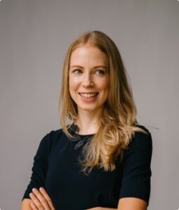 Smiling blonde woman in black dress with arms crossed.