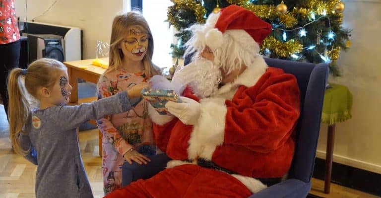 Two girls with face paint giving a gift to Santa Claus at Breakfast with Signing Santa event.