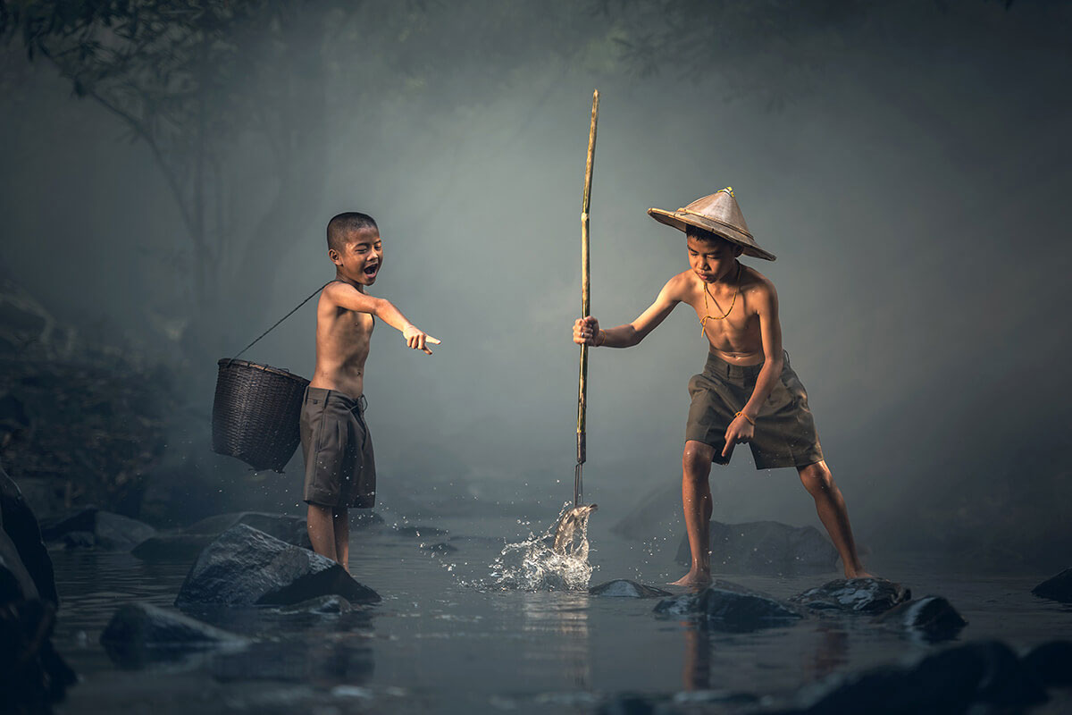 Two boys fishing in a river, one with a traditional hat, the other with a basket.