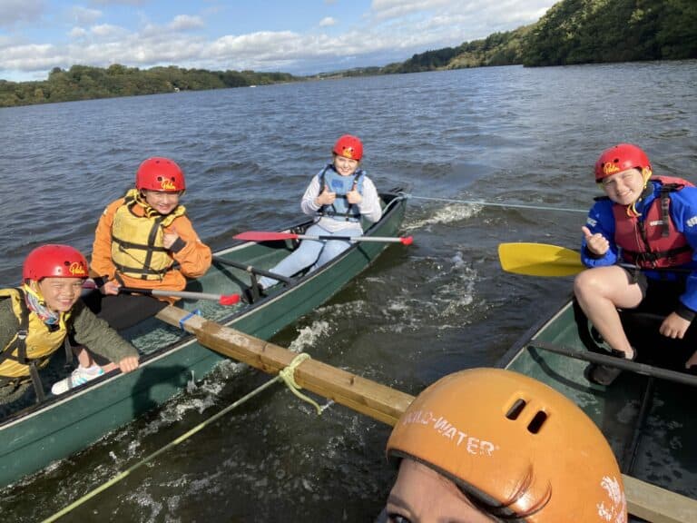 Four kids in canoes connected by rope, wearing helmets and life jackets, giving thumbs up on a lake.