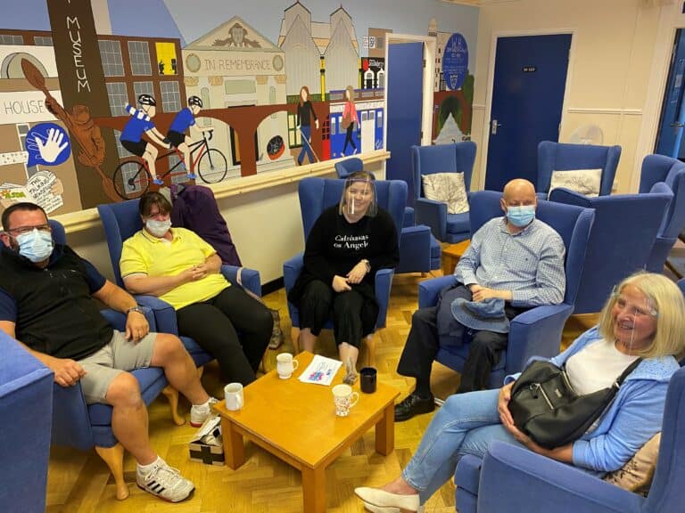 Group of people in blue chairs, wearing masks and face shields, gathered around a table.