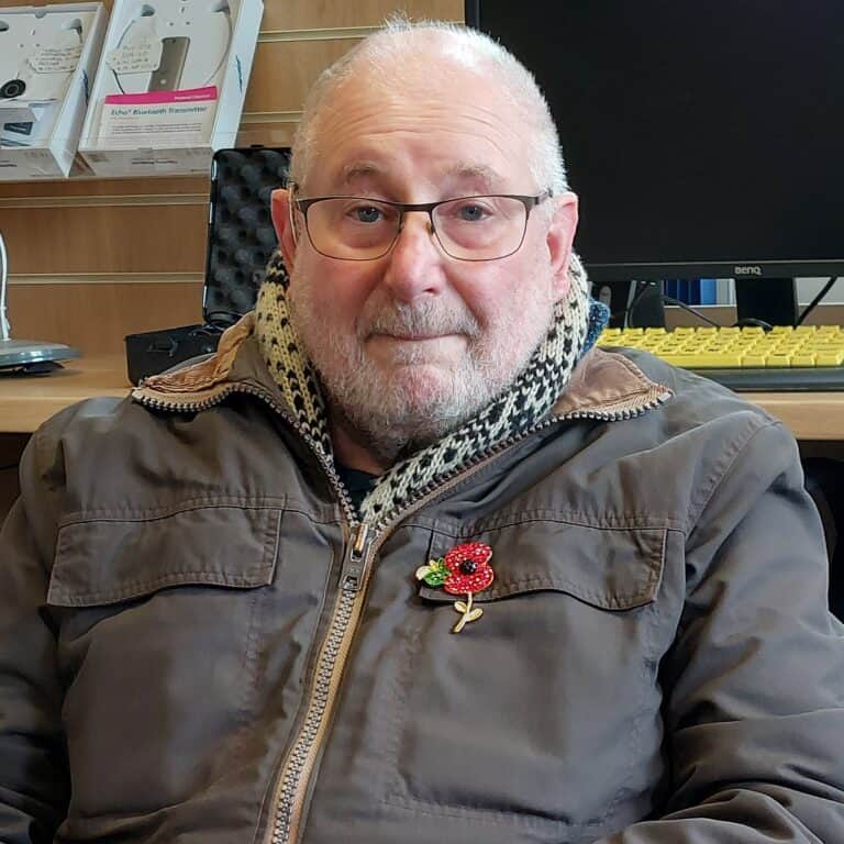 Man in jacket with poppy pin, seated at desk with computer. Adult Hearing Services.