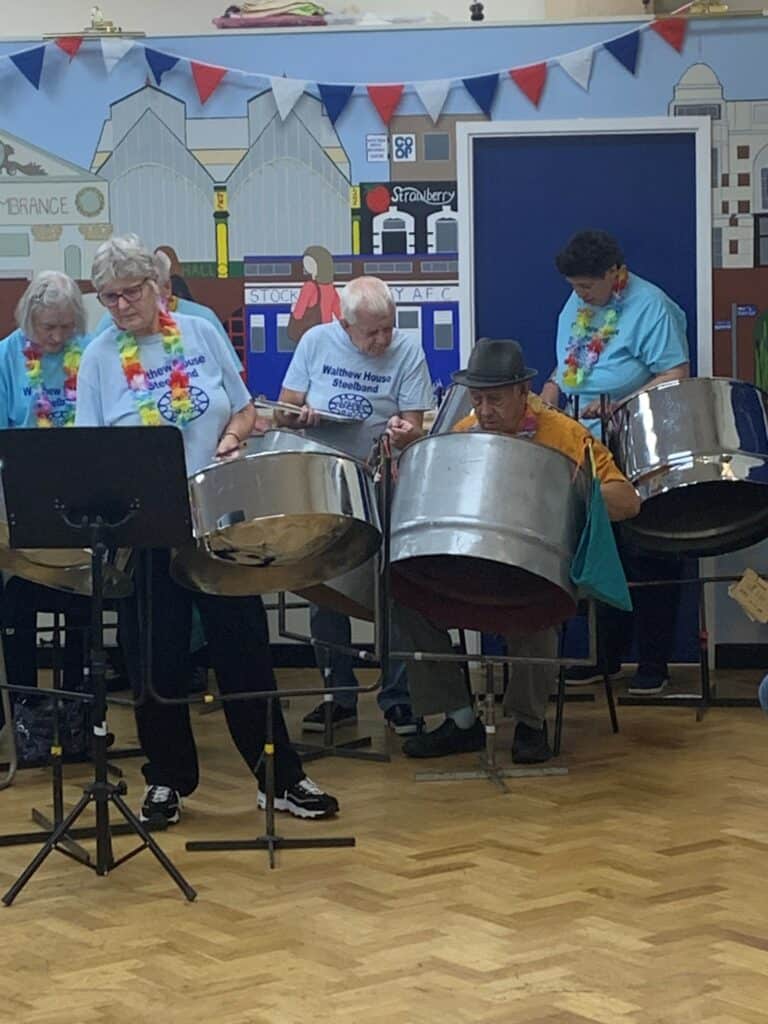 A group of men and women at the Proms wearing light blue t-shirts that say 'Walthew House Steelband.' They are wearing flower garlands and playing the steel drums, either standing or seated.