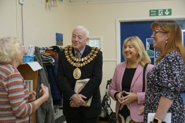 Walthew House: Mayor visits with two women, wearing mayoral chain and suit, pink jacket, and floral dress.