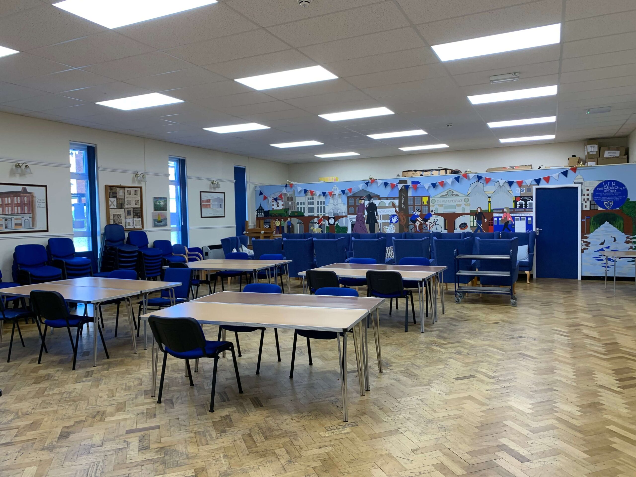 Empty community hall with tables, chairs, and a historical mural depicting local scenes.