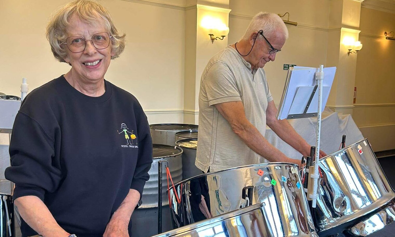 Two people playing steelpan drums, smiling at the camera.