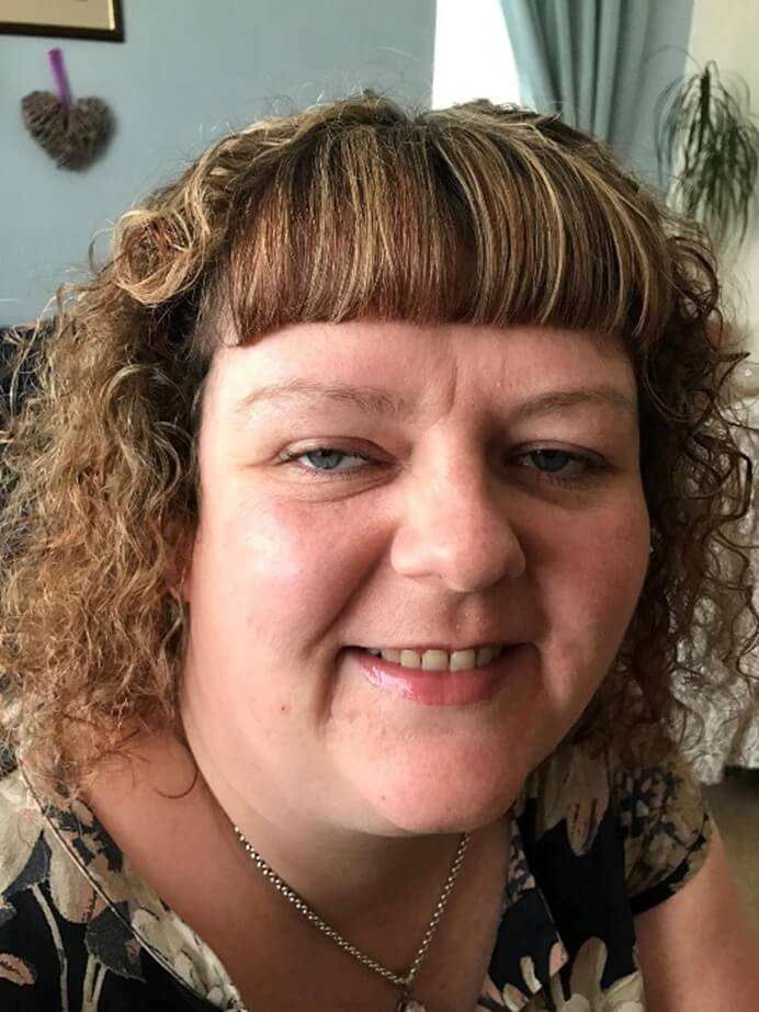 A photo of out chair, Rachael who is a woman with curly light brown hair and a fringe, smiling at the camera indoors. She is wearing a floral top and a silver necklace, with a heart-shaped wall decoration and curtains visible in the background.