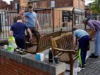 People restoring wooden park benches, with tools and drinks nearby.