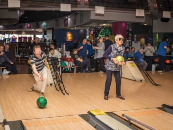 Woman with dark glasses bowling, man throwing a green ball. Blind Bowling.