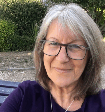 Mature woman with gray hair and glasses smiles outdoors with greenery in the background.
