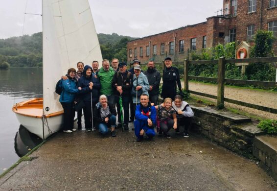 Group of people posing on a dock next to a sailboat by a lake, with a brick building in the background.