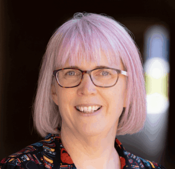 Sandi Marshall Smiling woman with pink hair and glasses wearing a colorful patterned shirt.