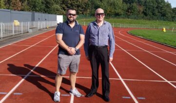 Two men stand on a running track, ready to race. One man wears a dark shirt and shorts; the other wears a light shirt and dark pants. Two men stand on a running track, ready to race. One man wears a dark shirt and shorts; the other wears a light shirt and dark pants.