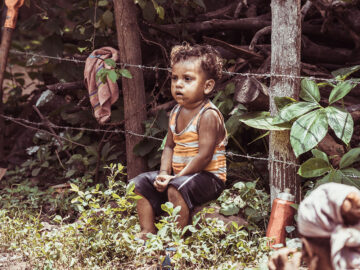 Child sits near a barbed wire fence in an outdoor setting.