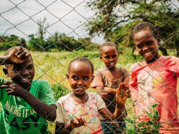 Four smiling African children standing behind a wire fence in a grassy field.