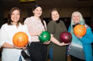 Four women smiling and holding bowling balls at a bowling alley, ready for a fun game.