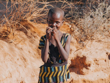 Young girl in patterned dress standing in a dry, sandy landscape.